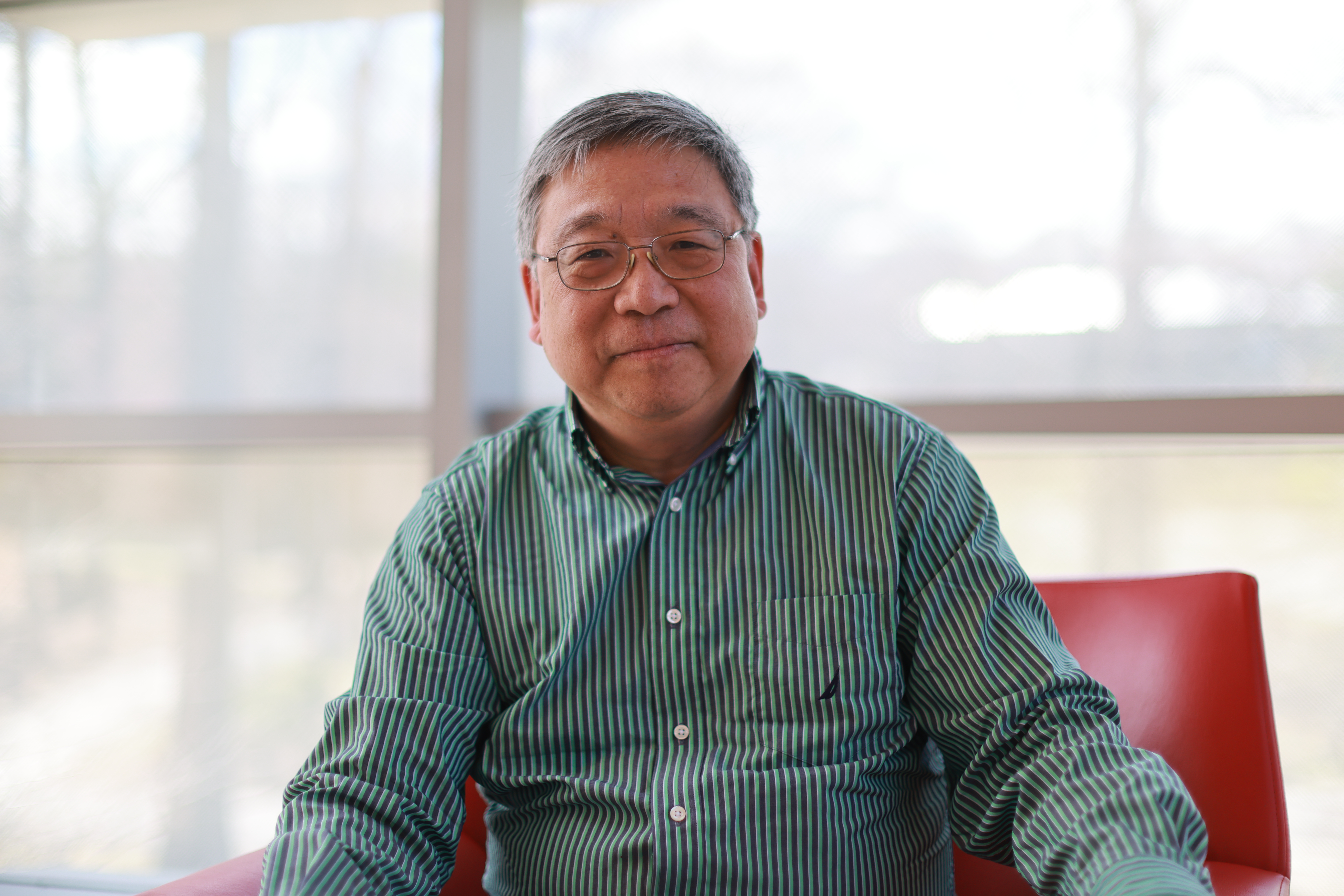 Professor Hong Qin, seated in a red chair in front of large windows