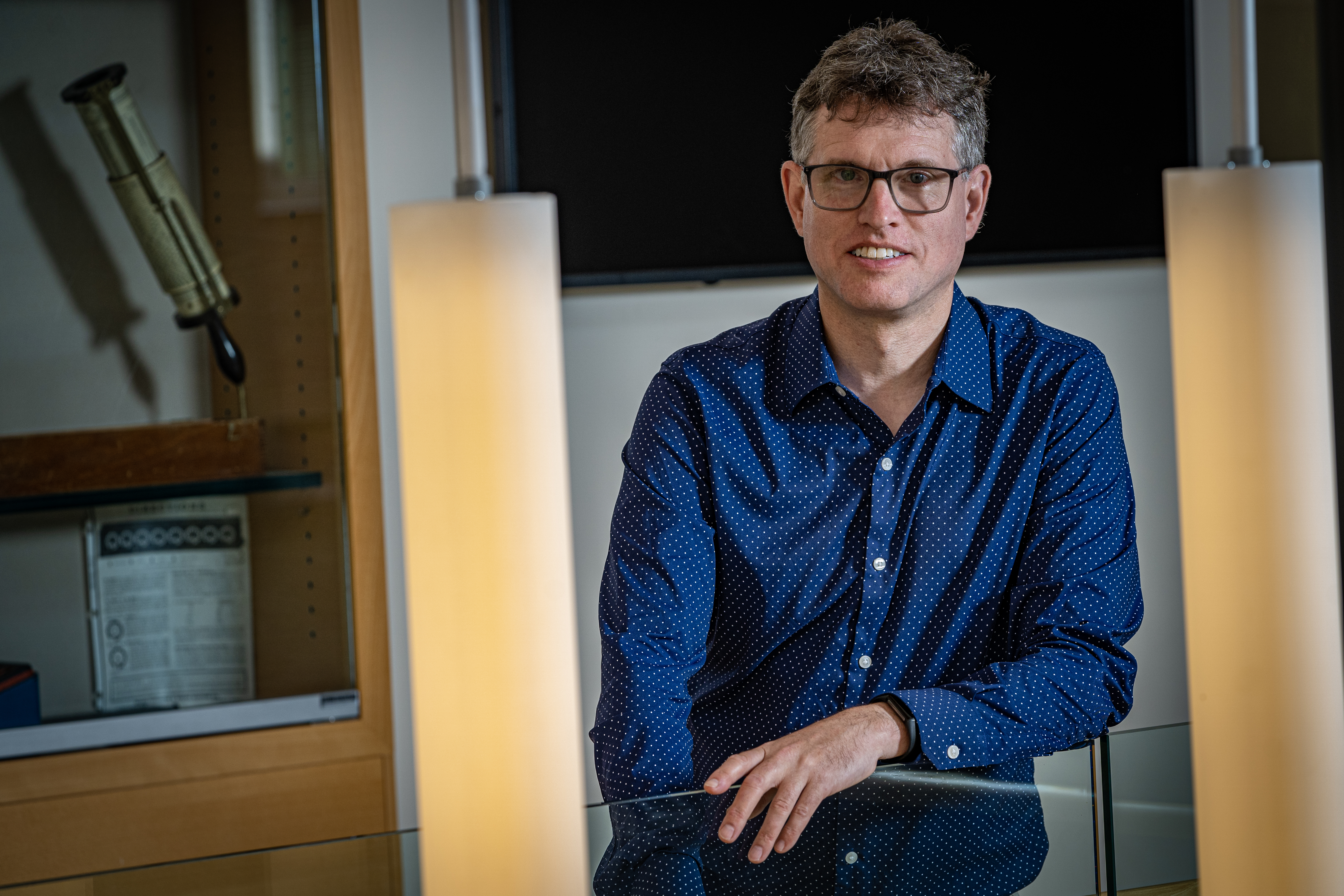 Michael Bender poses slightly behind hanging pendant lights, wearing a blue collared shirt and glasses.