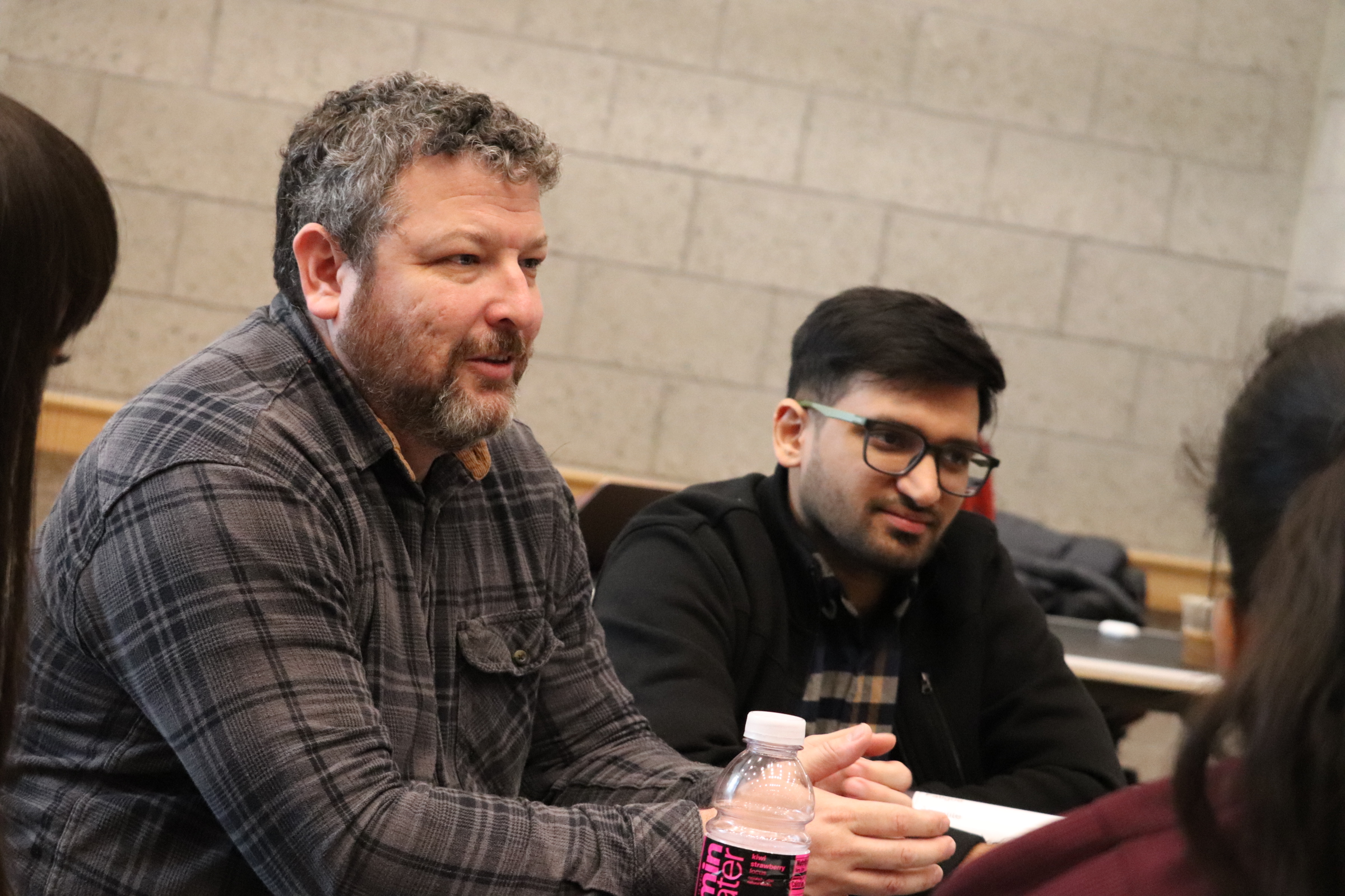 Man with graying hair in a gray checkered shirt sits at a table talking to students.