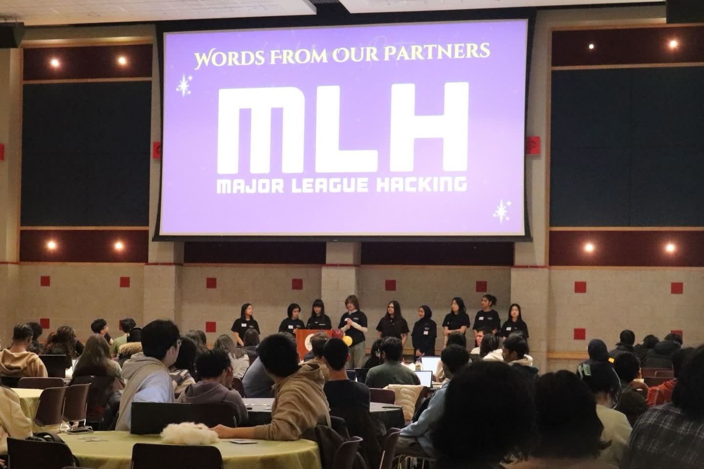 A wide-angle view of the ballroom where the event was hosted, showing the stage and the large projector overhead, which features the logo of the hackathon's sponsor