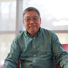 Professor Hong Qin, seated in a red chair in front of large windows