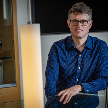 Michael Bender poses slightly behind hanging pendant lights, wearing a blue collared shirt and glasses.